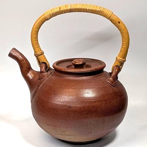 May include: A round, brown ceramic teapot with a textured, earthy finish. It features a curved spout, a matching lid with a small knob, and a tall, arched handle wrapped in light tan, natural-colored material attached to two side lugs.
