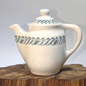 May include: Cream-colored ceramic teapot with a lid and handle. The teapot features a teal leaf and vine pattern around the body and on the lid. The teapot is sitting on a wooden surface.