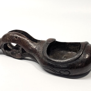 May include: A dark brown, antique-style, carved wooden vessel with a curved handle and a bowl-shaped body. The handle features an open design with decorative carvings. The vessel rests on a white surface.
