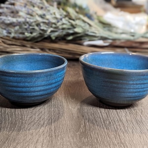 May include: Two small, handmade ceramic bowls with a textured, blue glaze. The bowls have a dark rim and sit on a wooden surface. The bowls are suitable for serving dips, snacks, or small portions.