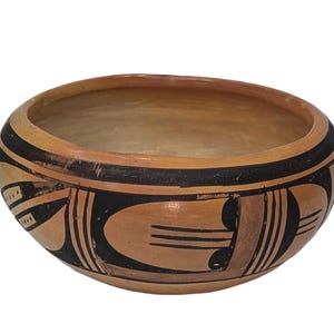 May include: A handmade, round pottery bowl with a light brown interior and exterior. The rim is decorated with a black band and geometric designs in black and tan. The bowl is likely used for serving or display.