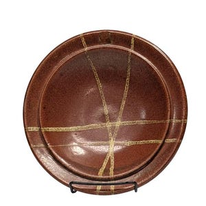 May include: A set of two brown ceramic plates with a textured surface and a network of light beige lines. The plates are displayed on a black metal stand, showcasing a rustic, handcrafted aesthetic.
