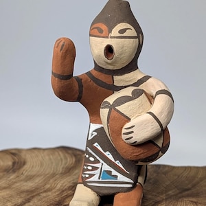 May include: A hand-painted ceramic figurine of a Native American figure. The figure is in a standing pose, with one arm raised and the other holding a round object. The figurine features brown, tan, and white colors with geometric patterns.
