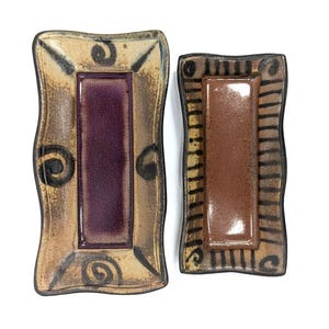 May include: Two ceramic rectangular trays with wavy edges. One tray has a tan border with a purple center and black swirl designs. The other tray has a brown border with a brown center and black stripe designs.