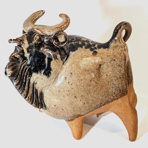 May include: A ceramic bull sculpture with a speckled tan body, dark blue and black accents, and tan legs and horns. The bull has a detailed face and a ring in its nose. The sculpture is a decorative item.