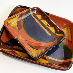 May include: Two square ceramic serving dishes. The larger dish is a reddish-orange color with a black rim. The smaller dish is nested inside. Both dishes have a colorful abstract design with yellow, blue, and green accents.