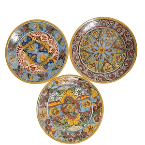 May include: Three decorative ceramic plates with intricate floral designs. Each plate features a unique pattern in shades of blue, yellow, brown, and green, with a gold-colored rim. The plates are arranged in a triangular formation against a white background.
