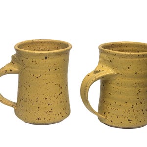 May include: Two cylindrical ceramic mugs with a speckled yellow glaze. Each mug features a curved handle attached to the side and a slightly flared rim. The surface of the mugs displays a textured appearance with dark brown speckles scattered across the yellow base color, creating a consistent look for both drinking vessels.