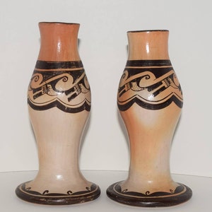 May include: Two ceramic vases with a light brown and beige color scheme. Each vase features a flared base, a slender body, and a wider top. The vases are adorned with black geometric patterns and wave-like designs, adding a touch of artistry.