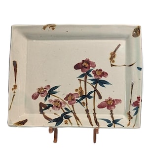 May include: A rectangular ceramic serving tray with a cream-colored base. The tray is decorated with a floral design featuring maroon flowers, green leaves, and brown stems. The tray is supported by a brown stand.