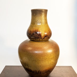 May include: A two-piece ceramic vase with a bulbous base and a smaller, rounded top. The vase has a gradient of colors, transitioning from a dark brown at the base to a lighter, golden-brown hue at the top. The vase sits on a dark wooden surface.