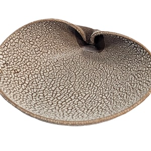 May include: A handcrafted, oval-shaped ceramic bowl with a unique, textured surface. The bowl features a cracked, off-white and brown pattern, with a folded rim. The design is organic and artistic, suitable for decorative or functional use.