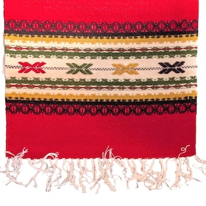 May include: A red woven textile with a cream-colored central band featuring embroidered cross and geometric patterns in red, green, yellow, and black. The bottom edge has white fringe.