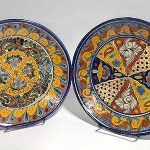May include: Two decorative ceramic plates with intricate hand-painted designs. The plates feature a vibrant color palette of blue, yellow, orange, and green. Each plate has a unique central pattern surrounded by a detailed border.
