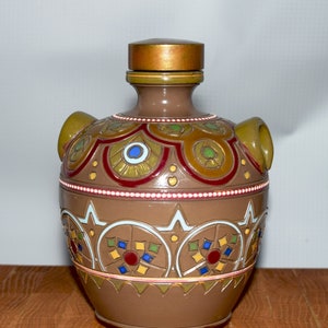 May include: A brown ceramic vase with a decorative design. The vase has a gold-colored lid and two handles. The design includes red, blue, green, and yellow accents. The vase is sitting on a wooden surface.