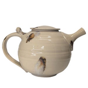 May include: A round ceramic teapot with a cream-colored glaze and a speckled finish. The teapot features a curved handle, a short spout, and a matching lid. Small, hand-painted brown and white bird illustrations are visible on the exterior surface.
