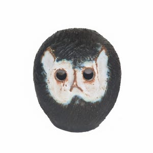 May include: A ceramic owl figurine with a textured, dark gray body and a white face. The owl has large, round eye sockets and a small beak. The face is outlined with a blueish-gray glaze.
