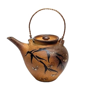 May include: A handcrafted ceramic teapot with a brown and tan glaze. The teapot features a black bamboo leaf design and a copper wire handle. The lid is brown and the spout is curved. The teapot is a unique piece of kitchenware.