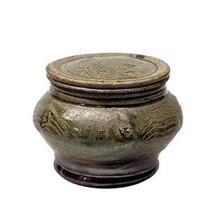 May include: A handcrafted ceramic jar with a lid. The jar is a muted green color with a dark brown base. The lid has a textured design. The jar has a round shape with carved details.