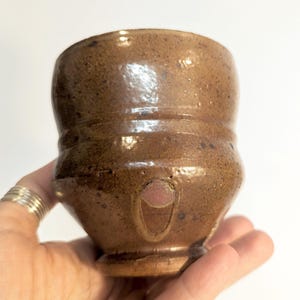 May include: A handmade brown ceramic cup with a unique shape. The cup has a textured surface with dark speckles and a small, decorative design. The cup is held in a hand against a white background. The cup has a small base.