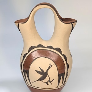 May include: Handmade pottery vase with a cream-colored body and a brown base, rim, and handle. The vase features a central design of a bird with a brown arch and black accents. The vase has a unique double-spout design.