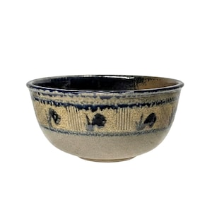 May include: A ceramic bowl with a wide rim and a rounded body. The bowl features a dark blue rim, a tan band with dark blue floral designs, and a light gray base. The bowl is suitable for serving food.