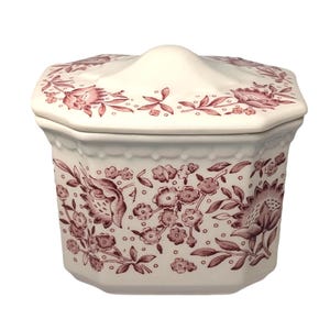May include: A white ceramic lidded jar with a floral pattern in burgundy. The jar has an octagonal shape with a slightly domed lid. The floral design includes flowers, leaves, and stems. The jar is suitable for storing small items.