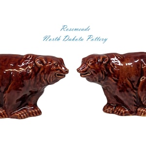 May include: Two brown ceramic bear figurines facing each other. The bears are glossy and detailed, with visible fur texture. The text "Rosemeade North Dakota Pottery" is above the bears.