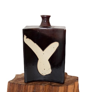 May include: A dark brown, rectangular ceramic bottle with a cream-colored, abstract brushstroke design. The bottle has a narrow neck and is displayed on a wooden base. The ceramic bottle is likely for decorative purposes.