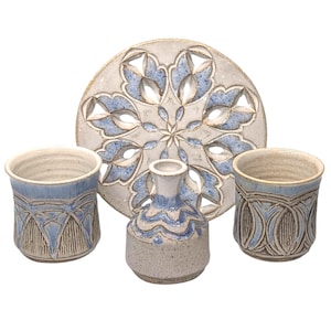 May include: A ceramic set featuring a large, round, decorative plate with a blue and white floral design, two matching cups, and a small vase. The pieces have a textured, handcrafted appearance.