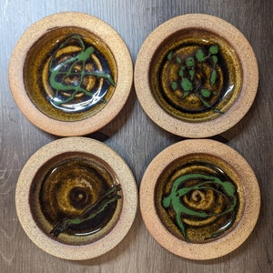 May include: Four handmade ceramic coasters. Each coaster features a circular design with a brown and green color scheme. The center of each coaster has a unique green abstract design, and the outer rim is a lighter brown.
