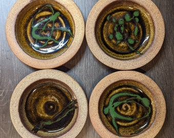 May include: Four handmade ceramic coasters. Each coaster features a circular design with a brown and green color scheme. The center of each coaster has a unique green abstract design, and the outer rim is a lighter brown.