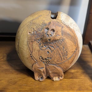 May include: A handcrafted, round ceramic vase with a textured, earthy appearance. The vase features a detailed, carved design on its surface, showcasing a blend of tan and brown hues. The vase has a small opening at the top and is set on a wooden surface.