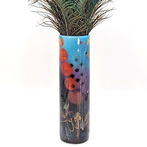 May include: A tall, cylindrical vase with a vibrant abstract design. The vase features a gradient of colors, transitioning from blue at the top to purple and black at the bottom. It is adorned with red circles, black squares, and gold accents. The vase is filled with peacock feathers.