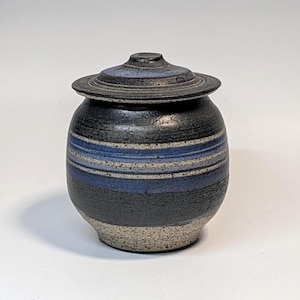 May include: A round ceramic jar with a matching lid, featuring a textured black and speckled gray base. The body is decorated with horizontal bands of deep blue and white stripes, creating a layered appearance on the dark, matte-finished surface.
