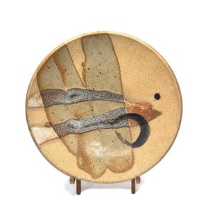 May include: A round ceramic plate with a unique design. The plate features a speckled tan background with abstract shapes in shades of brown, gray, and black. It is displayed on a small wooden stand.