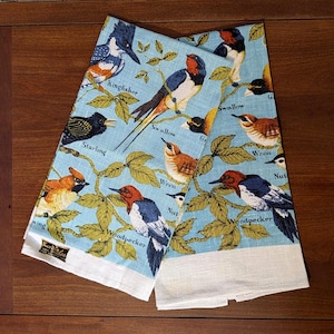 May include: Two light blue kitchen towels featuring a colorful bird pattern with illustrations of a Kingfisher, Swallow, Starling, Wren, and Woodpecker. Each bird is labeled with its name in black text, surrounded by green leaves and branches.