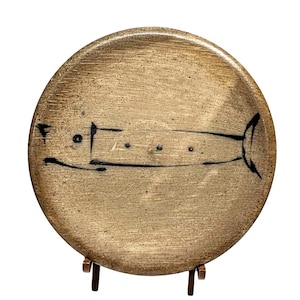 May include: A round, beige ceramic plate with a hand-drawn black fish design. The plate is displayed on a small, brown wooden stand. The fish illustration is simple, with a rectangular body and a detailed tail.