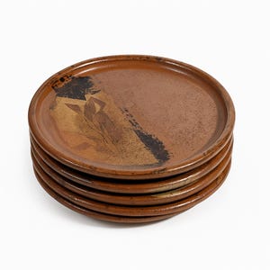 May include: A stack of six brown ceramic plates. Each plate has a circular shape with a raised rim. The plates have a decorative design in gold and black on the top surface.