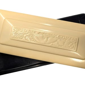 May include: A rectangular black plastic tray with a removable cream-colored lid. The lid features a raised, embossed central panel displaying a detailed relief pattern of a bird surrounded by stylized floral and leaf motifs across its surface.