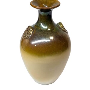 May include: A ceramic vase with a wide base and a narrow neck, featuring a gradient of colors from cream to brown to a dark bronze rim. The vase has two decorative shell-shaped accents on the sides.