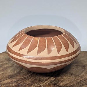 May include: A round, open-mouthed ceramic bowl with a reddish-brown base color and cream-colored geometric patterns. The bowl is resting on a wooden surface. The design features repeating triangular shapes around the rim.