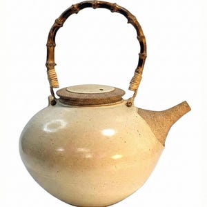 May include: Cream-colored ceramic teapot with a bamboo handle and a matching lid. The spout and lid are a darker, textured brown. The handle is made of curved bamboo. The teapot has a rounded body and a minimalist design.