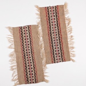 May include: Two rectangular beige woven placemats with a central patterned design. The design features vertical stripes of pink, black, and white geometric shapes. Each placemat has a fringed edge.