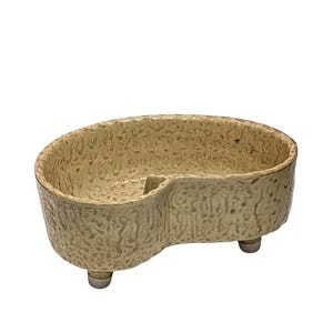 May include: A beige ceramic dish with a textured surface and a kidney bean shape. The dish has two compartments and rests on three small, rounded feet. The overall design is simple and functional.