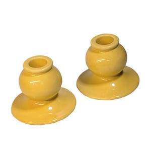 May include: Two yellow ceramic candle holders. Each holder has a round base with a small, spherical top that has a circular opening. The candle holders are a solid, glossy yellow color and are set against a white background.