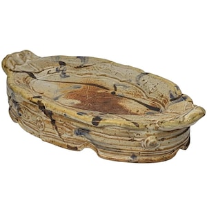 May include: A handmade ceramic soap dish with a unique, organic shape. The dish features a blend of beige, brown, and black colors, with a textured surface and decorative edges. The dish is approximately 8 inches long.
