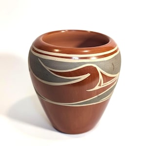 May include: A terracotta-colored ceramic vase with a rounded top and a tapered base. The vase features a decorative band of gray, white, and red abstract designs around the middle. The interior of the vase is a darker shade of terracotta.