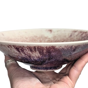 May include: A handmade ceramic bowl with a wide, shallow design. The bowl features a speckled, mottled glaze in shades of purple, burgundy, and cream. The bowl is held in a hand, showcasing its shape and color variations.
