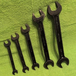 May include: A set of five black metal open-ended wrenches arranged in a diagonal line from smallest to largest. Each tool features a double-ended design with an open jaw at both ends, showing a dark, matte finish and visible stamped text markings.